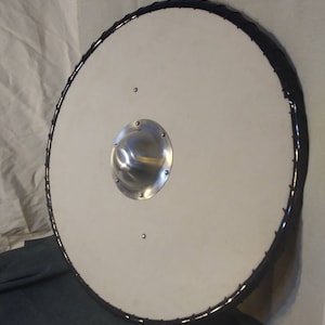 May include: A white round shield with a black leather trim and a metal boss in the center. The shield is made of wood and has a diameter of approximately 24 inches.