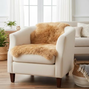May include: A cream-colored armchair with dark wooden legs, featuring a tan sheepskin throw draped over the seat and back. The room has a neutral color palette, with plants and a sofa visible in the background.
