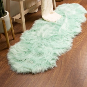 May include: A long, fluffy, mint green faux fur rug. The rug is rectangular with a rounded shape and is placed on a wooden floor. The rug is a home decor item.