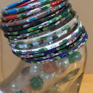 May include: A stack of enamel bangles in various colours, including red, blue, green, and white, resting on a clear glass bottle. The bangles feature floral and abstract designs. The bottle has a silver cap and is filled with decorative stones.
