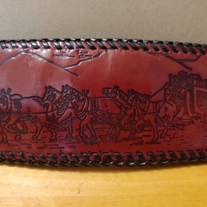 May include: A red leather wallet with a black stitched border. The wallet has an embossed design of a horse-drawn carriage with horses pulling it.