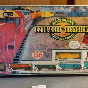 Puede incluir: Una caja de set de tren a escala HO Bachmann E-Z Track Thunderbolt con una locomotora roja numerada 6067, un vagón cisterna amarillo y varios vagones de carga. El empaque muestra el texto 156 pieces e ilustraciones del set.