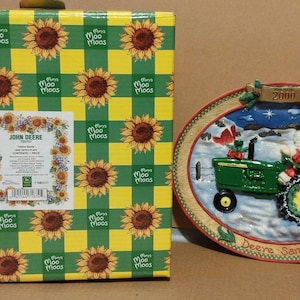 Könnte beinhalten: Dekorative ovale Platte mit einem grünen John Deere Traktor und einer Weihnachtsmannfigur, datiert 2000. Die Schachtel mit einem Sonnenblumenmuster in Gelb und Grün ist ebenfalls sichtbar.