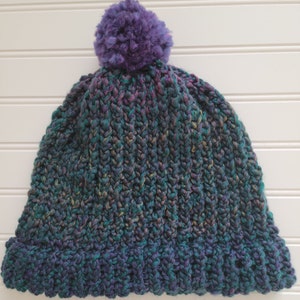 May include: A hand-knitted beanie hat in shades of teal, purple, and green with a large, fluffy purple pom-pom on top. The hat features a ribbed brim and a textured knit pattern, perfect for cold weather.