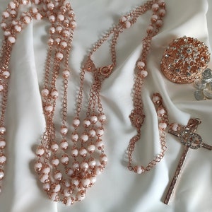 Può includere: Un rosario in oro rosa con perline bianche e una croce in oro rosa. Il rosario è su uno sfondo di tessuto bianco. C'è una piccola scatola in oro rosa con strass e alcune monete d'argento sullo sfondo.