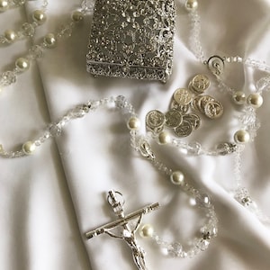 Puede incluir: Un rosario de plata con cuentas de cristal transparente y detalles de perlas blancas. El rosario está colocado sobre un fondo de tela blanca con una caja de plata y monedas de plata.