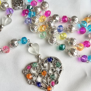 May include: A colorful beaded necklace with a silver heart pendant. The necklace features a variety of faceted beads in shades of pink, blue, yellow, and green. The heart pendant is silver with colorful beads. The necklace is on a white background.