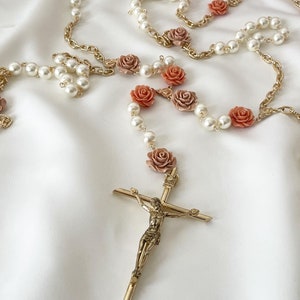 Può includere: Un rosario con perline di finta perla, catena color oro e accenti di rose corallo e malva. Il rosario presenta un crocifisso color oro con una dettagliata raffigurazione di Gesù. Lo sfondo è un tessuto bianco e serico.