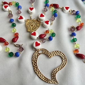 May include: A collection of colorful necklaces with heart-shaped pendants and beads. The necklaces feature white beads with red hearts, gold-colored heart pendants, and a variety of colorful beads in red, green, blue, and purple.