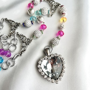 May include: A silver-toned necklace with a heart-shaped pendant. The pendant is encrusted with clear crystals and surrounded by smaller stones. The necklace features a chain of colorful beads in shades of pink, blue, and yellow.
