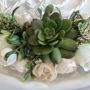 Può includere: Una composizione floreale con una grande succulenta verde al centro, circondata da rose bianche e color crema e verde. La composizione è accentuata da piccoli fiori bianchi e un cordoncino bianco intrecciato.