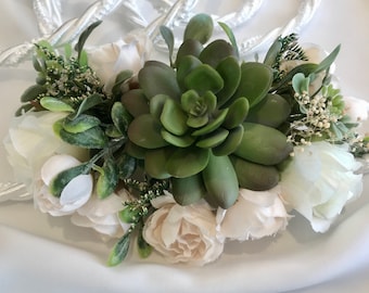 Puede incluir: Un arreglo floral con una gran suculenta verde en el centro, rodeado de rosas blancas y crema y vegetación. El arreglo se acentúa con pequeñas flores blancas y un cordón blanco trenzado.