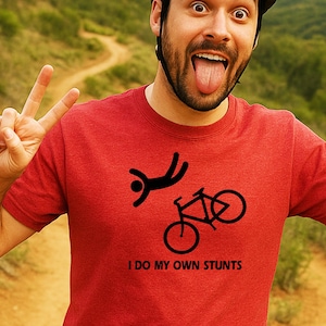 May include: Red t-shirt with a black graphic of a person falling and a bicycle. The text "I DO MY OWN STUNTS" is printed below the graphic. The person in the image is wearing a helmet and making a funny face.