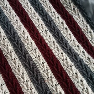 May include: A crocheted blanket with stripes of burgundy, white, and gray yarn. The blanket has a cable knit pattern.