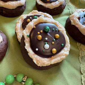 Peut inclure: Gros plan sur plusieurs biscuits au chocolat avec une base de glaçage de couleur claire et une garniture au chocolat noir. Les garnitures sont décorées de paillettes vertes, dorées et blanches. Un collier de perles vertes est au premier plan.