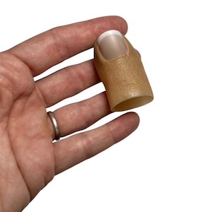 May include: A prosthetic thumb tip cover featuring a realistic skin-toned silicone texture and a white-tipped fingernail. The object is designed to fit over a human thumb, providing a natural appearance with detailed skin texture and a French manicure style.