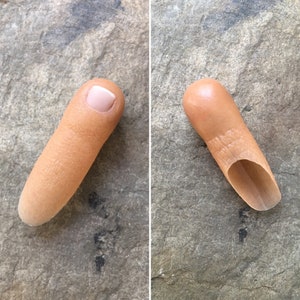 May include: Two close-up images of a prosthetic fingertip. The first image shows the fingertip from the side, with a nail painted a light pink colour. The second image shows the fingertip from the top, with a hollowed-out opening.