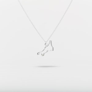 Puede incluir: Un collar de plata con un colgante con forma de costa. El colgante es un diseño minimalista, un fino contorno de la masa terrestre suspendido de una delicada cadena. El collar se muestra sobre un fondo blanco.