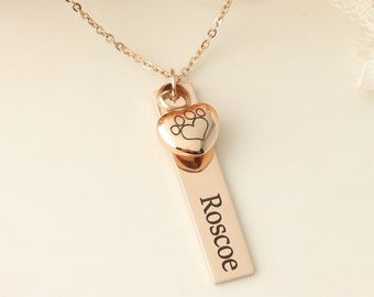 May include: Rose gold necklace with a heart-shaped pendant featuring a paw print design. A rectangular tag with the name "Roscoe" is attached. The necklace is displayed against a soft, floral background.