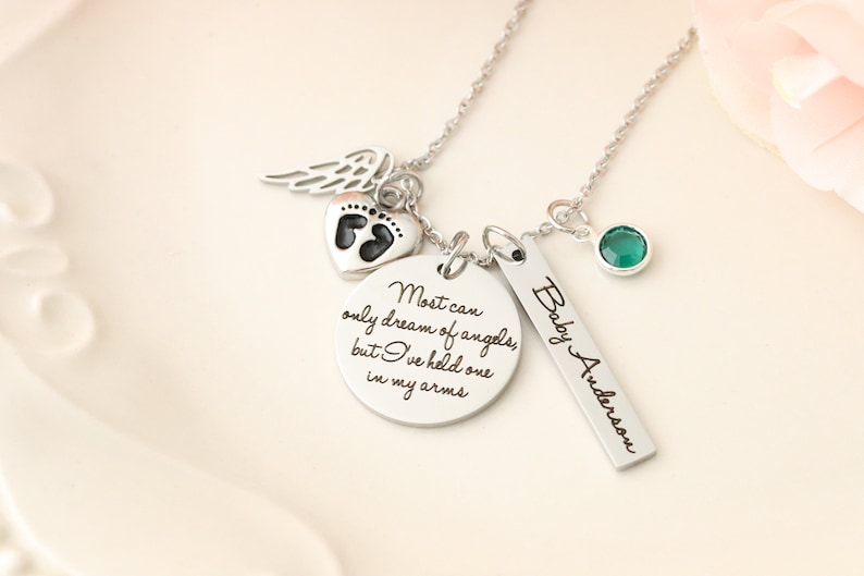 May include: Silver necklace with a charm that reads "Most can only dream of angels, but I've held one in my arms."  The necklace also features a small heart charm with a footprint, a small angel wing charm, and a green gemstone charm.  The necklace also has a tag with the text "Baby Anderson".