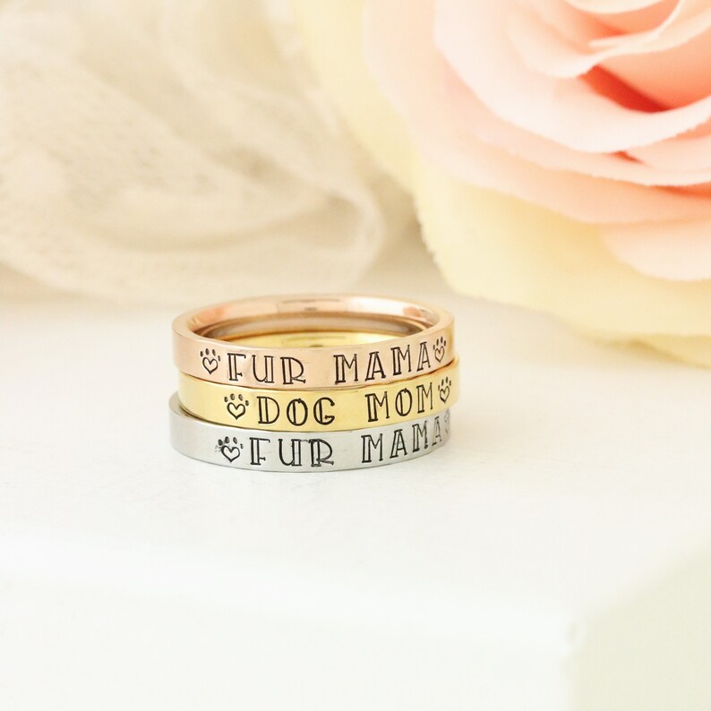 Puede incluir: Tres anillos apilados uno encima del otro en oro, plata y oro rosa. Los anillos est&aacute;n grabados con las palabras "Fur Mama" y "Dog Mom" con s&iacute;mbolos de huella de pata y coraz&oacute;n.