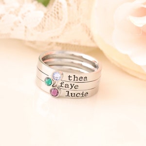 May include: A silver ring with three bands, each engraved with a name: Thea, Faye, and Lucie. Each band has a small gemstone: light blue, green, and purple.