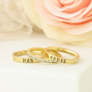 Puede incluir: Tres anillos de oro con una fila de peque&ntilde;as piedras transparentes en un anillo. Los anillos est&aacute;n grabados con los nombres "Max" y "Sadie".
