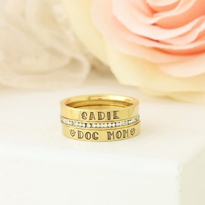 Puede incluir: Conjunto de tres anillos apilables de color dorado con las palabras "SADIE" y "DOG MOM" grabadas. El anillo del medio tiene una fila de piedras de circonita c&uacute;bica transparentes.