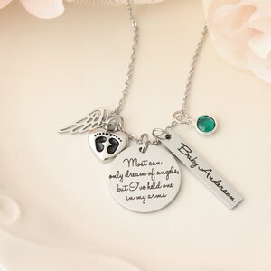 May include: Silver necklace with multiple charms, including a heart with footprints, a wing, a round charm with the text "Most can only dream of angels, but I've held one in my arms", a bar charm with the text "Baby Anderson", and a green gemstone.