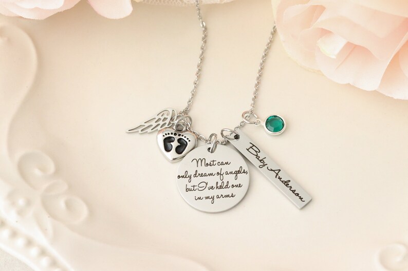 May include: Silver necklace with multiple charms, including a heart with footprints, a wing, a round charm with the text "Most can only dream of angels, but I've held one in my arms", a bar charm with the text "Baby Anderson", and a green gemstone.