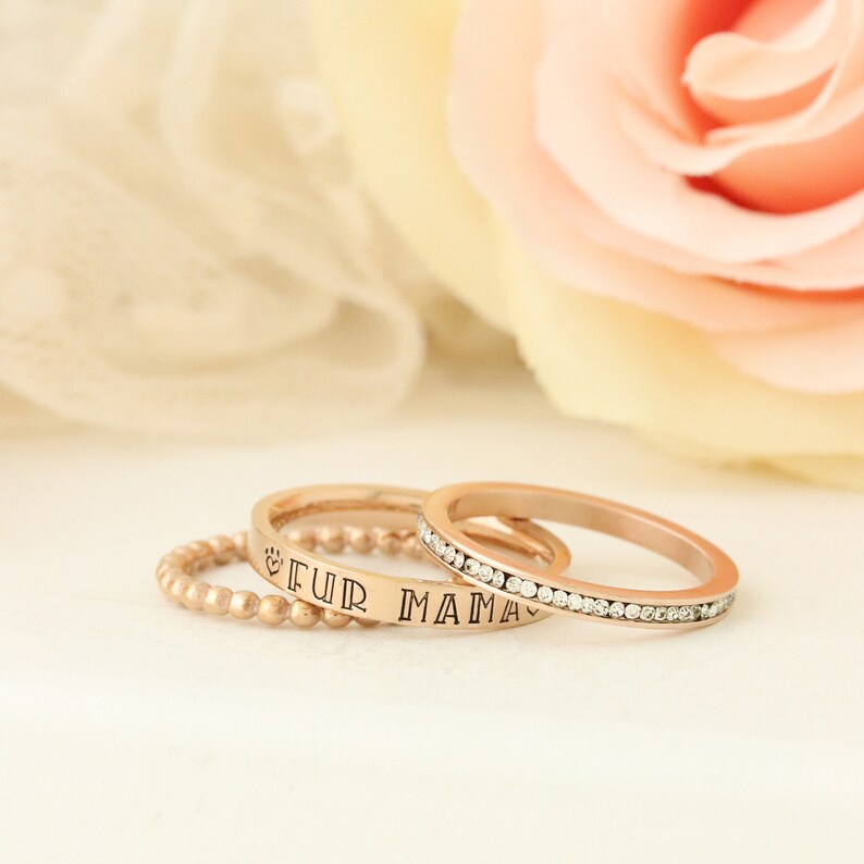Puede incluir: Tres anillos dorados apilados sobre una superficie blanca. El anillo superior tiene una fila de peque&ntilde;as piedras transparentes. El anillo del medio tiene las palabras "Fur Mama" grabadas. El anillo inferior es un anillo de cuentas.
