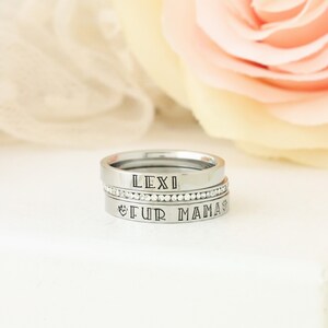 Puede incluir: Tres anillos de plata apilados uno encima del otro. El anillo superior dice "LEXI" en may&uacute;sculas. El anillo del medio es una banda de peque&ntilde;os diamantes de imitaci&oacute;n transparentes. El anillo inferior dice "FUR MAMA" en may&uacute;sculas con un s&iacute;mbolo de huella de pata a cada lado del texto.