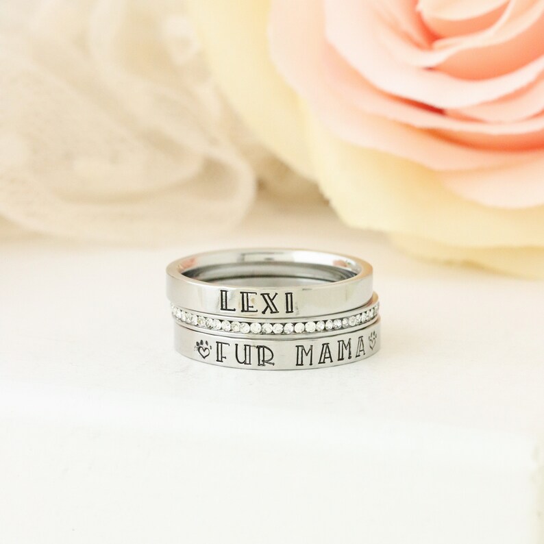 Puede incluir: Tres anillos de plata apilados uno encima del otro. El anillo superior dice "LEXI" en may&uacute;sculas. El anillo del medio es una banda de peque&ntilde;os diamantes de imitaci&oacute;n transparentes. El anillo inferior dice "FUR MAMA" en may&uacute;sculas con un s&iacute;mbolo de huella de pata a cada lado del texto.