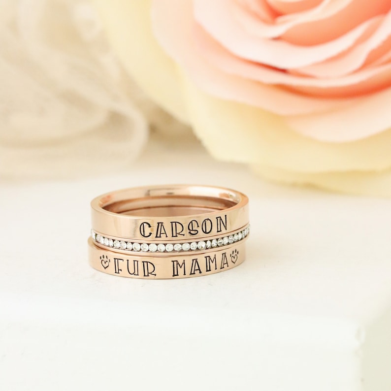Puede incluir: Anillos apilables de oro rosa con las palabras "CARSON" y "FUR MAMA" grabadas. El anillo superior tiene una fila de piedras de circonita c&uacute;bica transparentes.