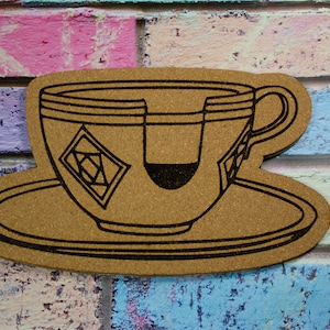 May include: A cork coaster shaped like a teacup and saucer with a black outline. The teacup has a tea bag inside and a black geometric design on the side.