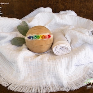 May include: A white, fringed, fabric backdrop with a wooden prop and a white fabric swaddle. The wooden prop has a rainbow-coloured headband attached to it.