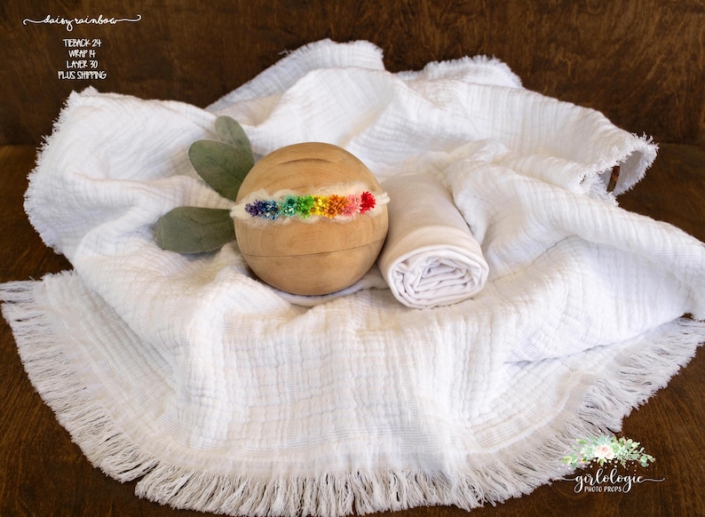 May include: A white, fringed, fabric backdrop with a wooden prop and a white fabric swaddle. The wooden prop has a rainbow-coloured headband attached to it.