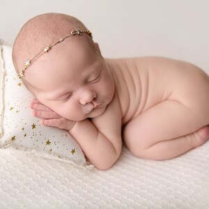 May include: A newborn baby sleeping on a white pillow with gold stars. The baby is wearing a gold headband with small stars.
