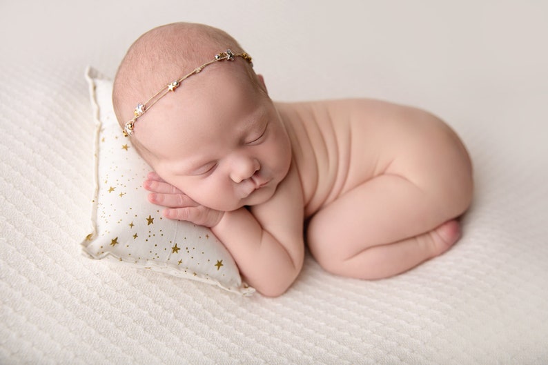 May include: A newborn baby sleeping on a white pillow with gold stars. The baby is wearing a gold headband with small stars.