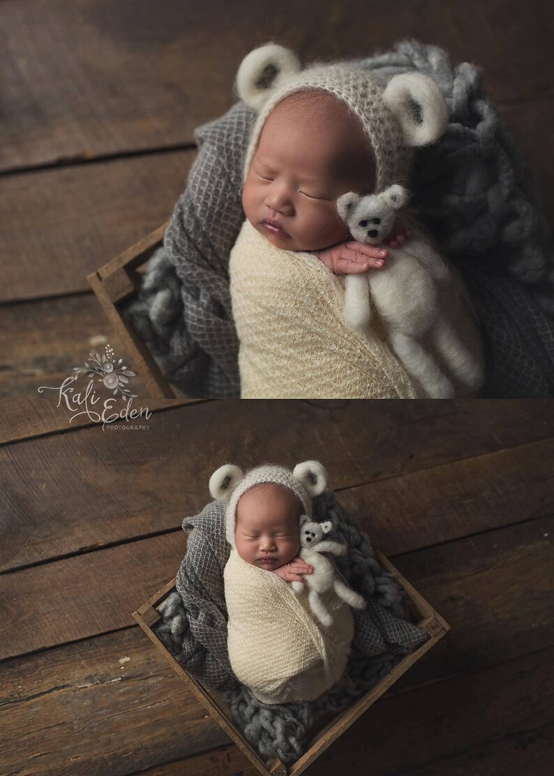 May include: A newborn baby wearing a white knitted bear hat and a white knitted blanket, sleeping in a wooden crate with a white knitted bear toy. The baby is surrounded by a gray knitted blanket.