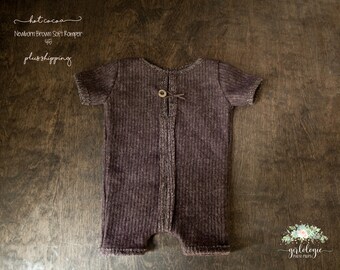 May include: A dark brown ribbed knit romper for newborns featuring short sleeves and a single wooden button closure at the center of the neckline. The garment has a textured fabric finish and a vertical ribbed pattern throughout the entire piece.