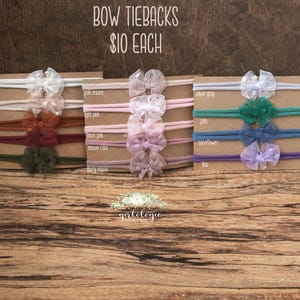 Könnte beinhalten: Mehrere Baby-Haarbänder mit Schleifen in verschiedenen Farben, darunter Off-White, Creme, Rost und Olive. Die Haarbänder werden auf Pappe präsentiert, mit dem Text "BOW TIEBACKS $10 EACH".