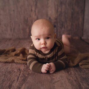 May include: A baby in a brown and green striped onesie is lying on its stomach on a brown wooden floor. The baby is looking directly at the camera with a serious expression.