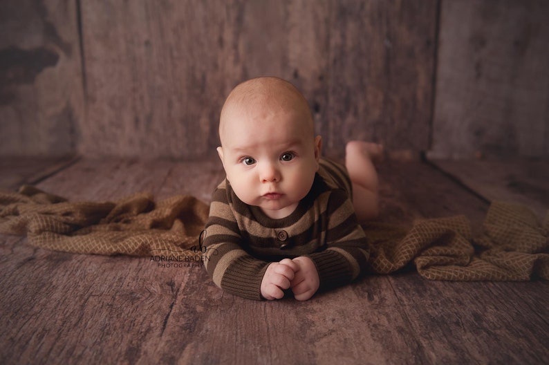 May include: A baby in a brown and green striped onesie is lying on its stomach on a brown wooden floor. The baby is looking directly at the camera with a serious expression.