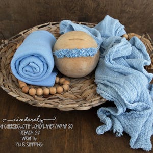 May include: A baby photography prop set in a woven basket. The set includes a rolled-up light blue wrap, a wooden head support with a blue headband, and draped cheesecloth fabric. A wooden bead garland and a decorative tassel are also included.