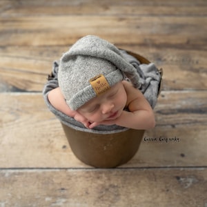May include: A newborn baby wearing a gray knitted hat with a brown leather patch that says "hello world" is sleeping in a brown bucket. The baby is wrapped in a gray blanket.