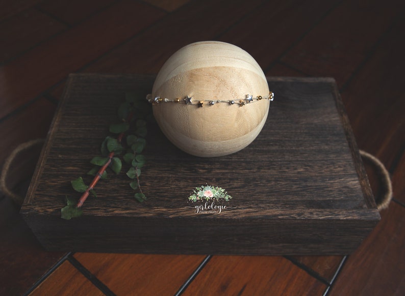 May include: A wooden ball with a gold and silver star headband on top of it. The ball is sitting on a dark brown wooden box with a floral logo that says "golelegie photography".