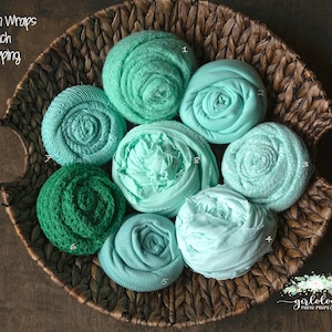 Op de afbeelding: Een rieten mand gevuld met acht stoffen swaddles in tinten mint en groen. De swaddles zijn gerold in een roosvorm. De tekst "Mint / Green Wraps $ 16 per stuk plus verzendkosten" is zichtbaar in de linkerbovenhoek van de afbeelding.
