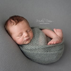 May include: A newborn baby is sleeping soundly, wrapped in a light green knitted blanket. The baby is lying on a gray background.