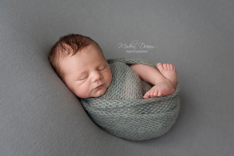 May include: A newborn baby is sleeping soundly, wrapped in a light green knitted blanket. The baby is lying on a gray background.